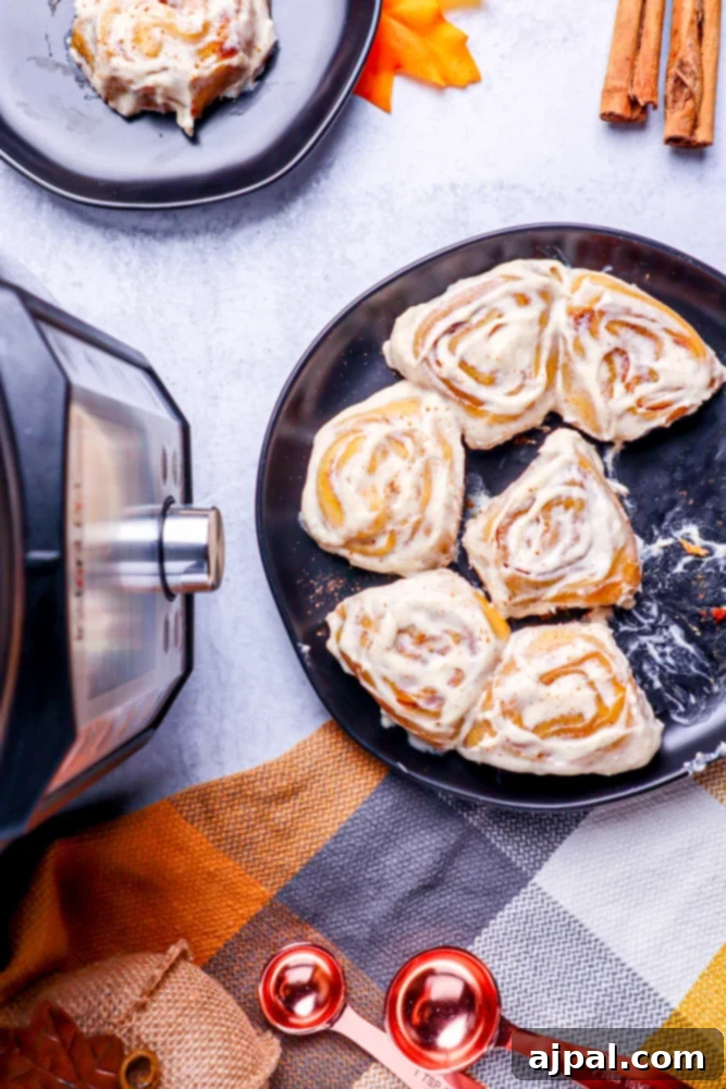 Easy Instant Pot Pumpkin Cinnamon Rolls 2 pumpkin cinnamon rolls in a black pan sitting next to a black and silver instant pot on a marble table with a checkered hand towel