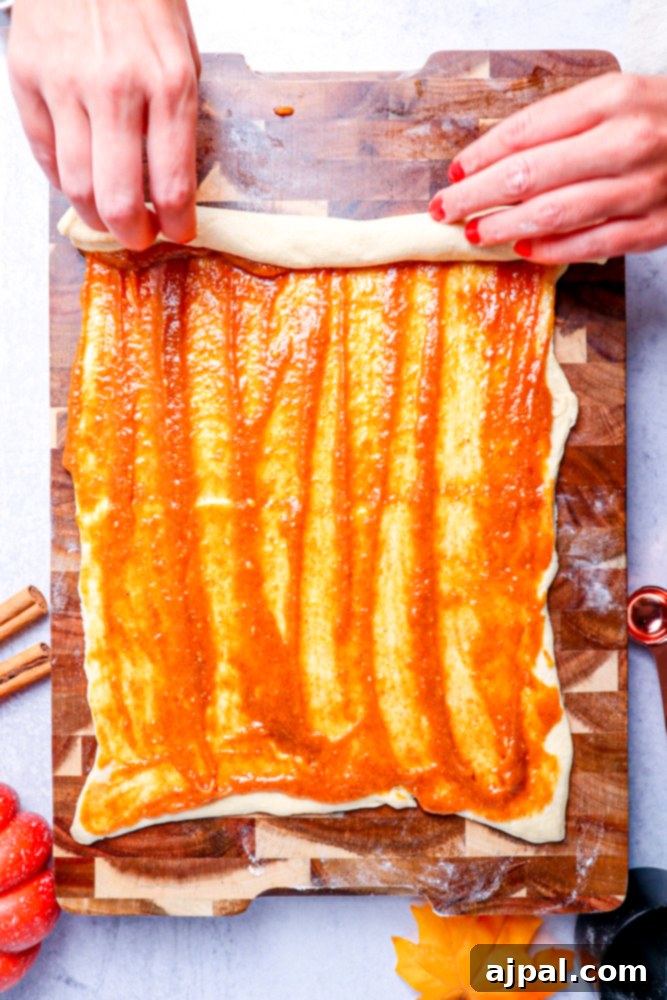 Pumpkin spice mixture spread across a flat sheet of dough on a wooden cutting board, with the dough in the process of being rolled up.