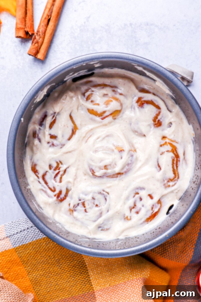 Eight pumpkin cinnamon rolls, nestled in a metal cake pan and generously blanketed with a thick layer of smooth white frosting.