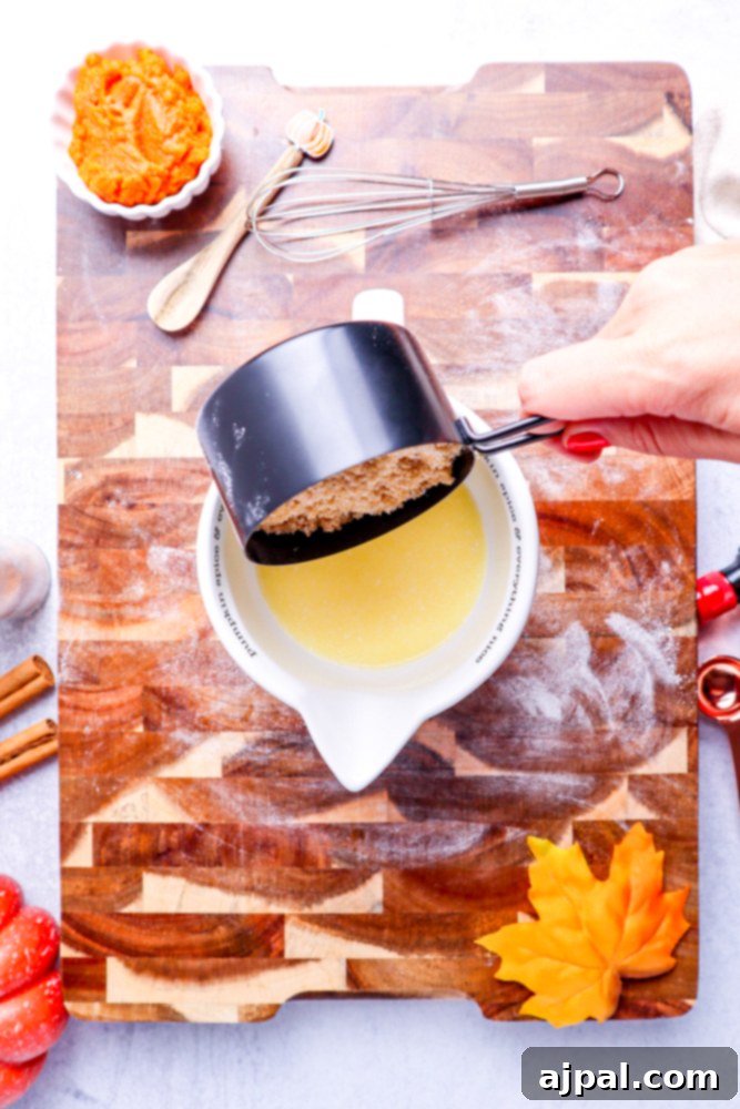 Brown sugar being carefully poured into melted butter in a white measuring cup, set against a wooden cutting board.