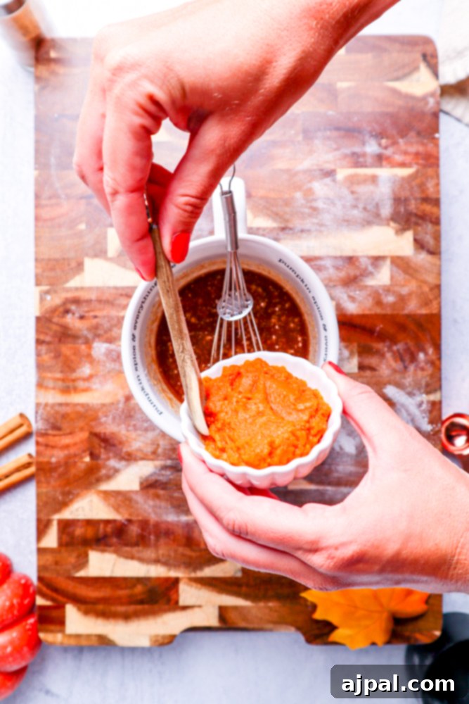 Spices being carefully added from a white bowl to a creamy mixture of melted butter and brown sugar in a white measuring cup on a wooden cutting board.
