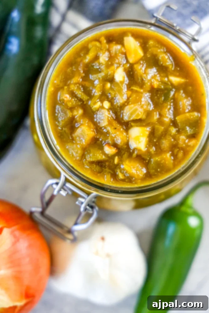 A jar of homemade green chile sauce, perfectly smooth and vibrant, with fresh green chile peppers and onions artfully arranged beside it, highlighting the key ingredients.