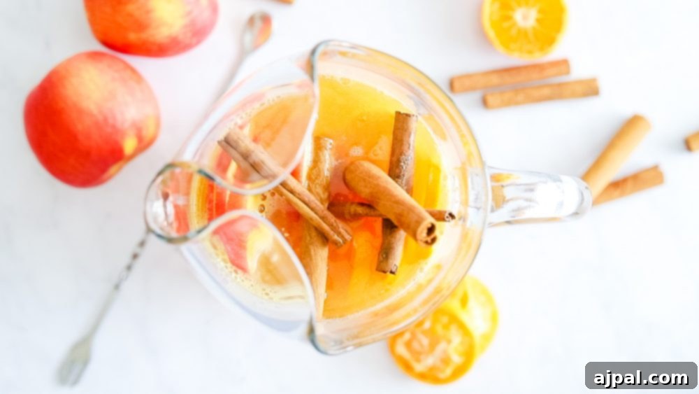 A large pitcher of amber-colored rum punch, generously garnished with thin slices of apple and orange, and several cinnamon sticks, ready to be poured for a gathering.