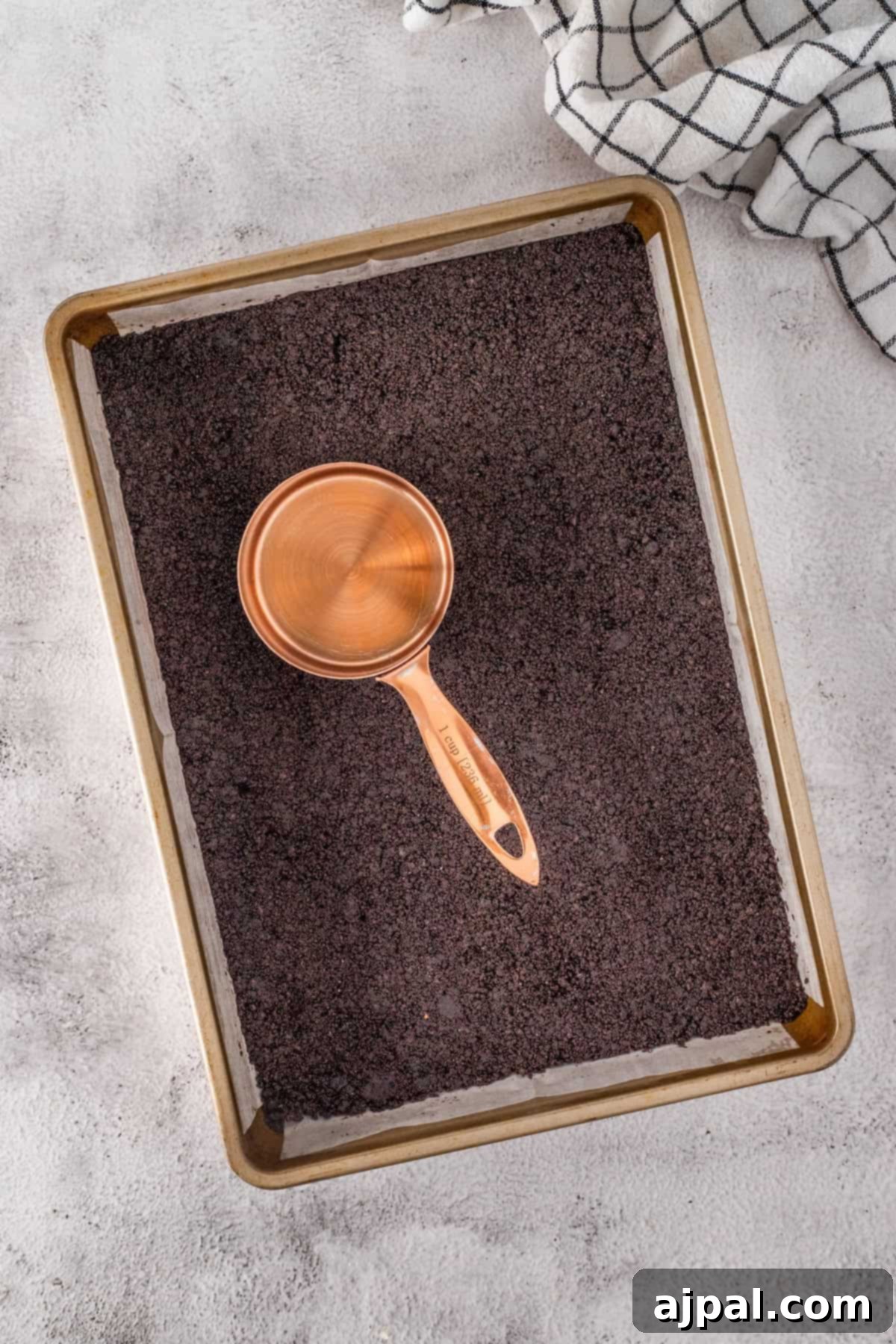 A copper measuring cup rests on a baking tray filled with finely ground dark material, reminiscent of cookie crumbs. A checkered cloth is visible in the top corner.