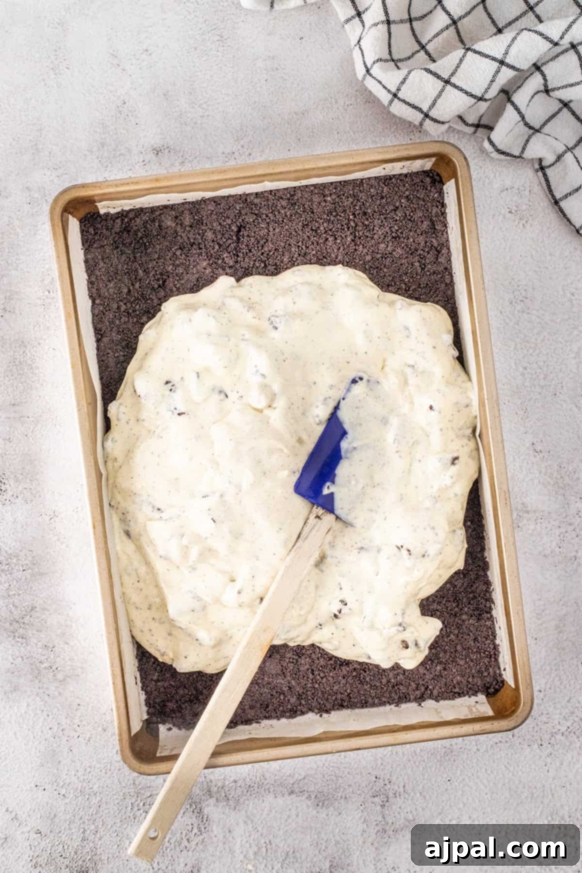 A baking tray holds a layer of crushed cookies beneath a rich spread of cream mixture, perfect for making cookies and cream cheesecake bars. A spatula rests on top, with a checkered cloth placed nearby.