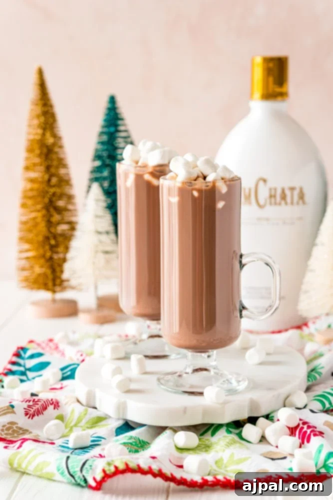 Creamy Rumchata Hot Chocolate Cocktail 2 Large, tall glasses of hot chocolate adorned with fluffy marshmallows, with a bottle of RumChata liquor in the softly blurred background.