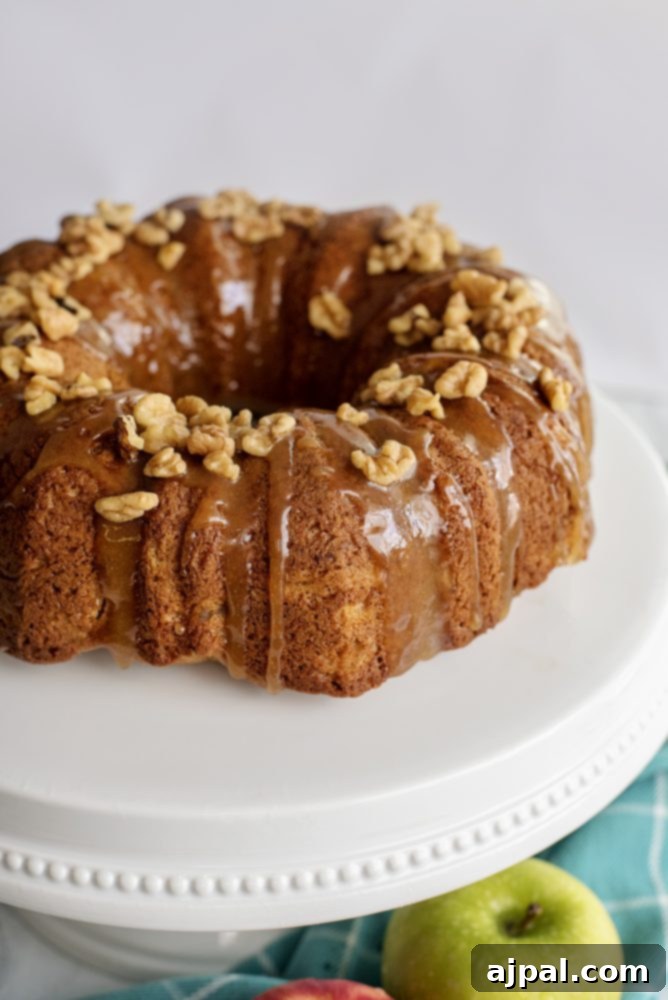 Decadent Caramel Apple Spice Cake Made Easy 11 A rich brown bundt cake adorned with a generous drizzle of caramel and a sprinkle of chopped walnuts and delicate apple slices, presented on a white plate.
