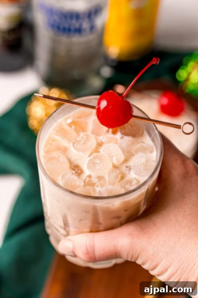 A perfectly chilled Irish Cream White Russian cocktail, garnished with a vibrant maraschino cherry on a toothpick, held in a hand against a soft background.