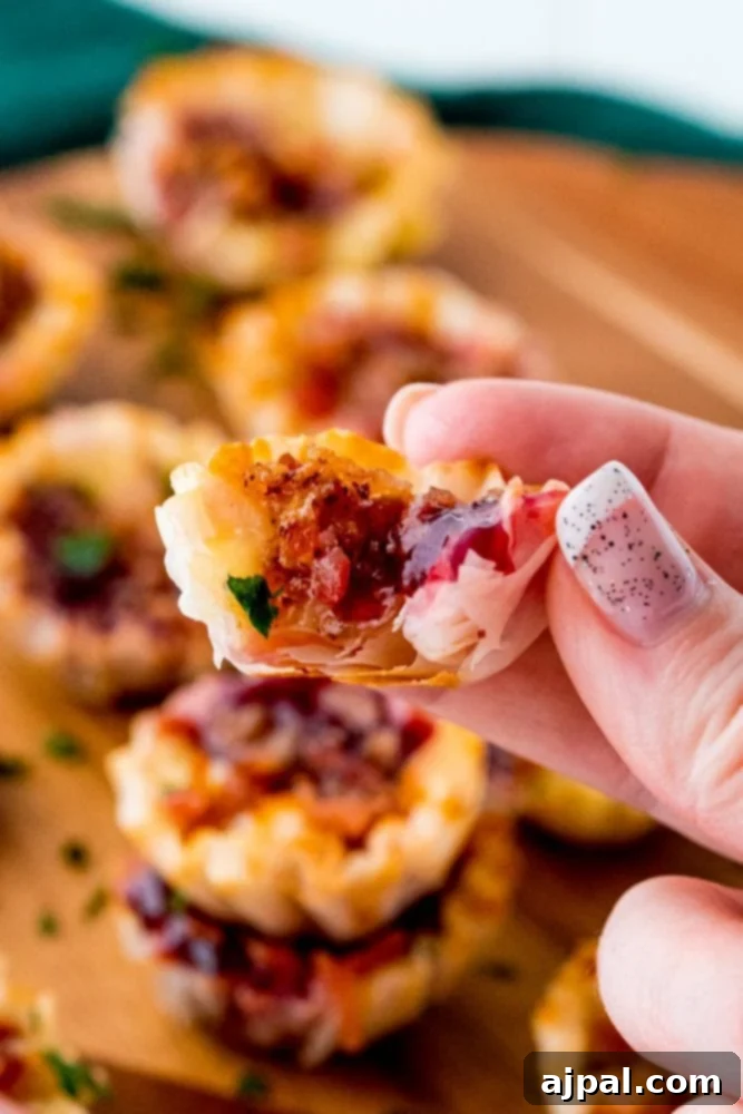 Cheesy Bacon Raspberry Morsels 12 A hand gracefully holds a small, perfectly golden phyllo cup, showcasing its filling of creamy cheese, sweet raspberry jam, and crispy bacon bits, highlighting the delectable layers of flavor.