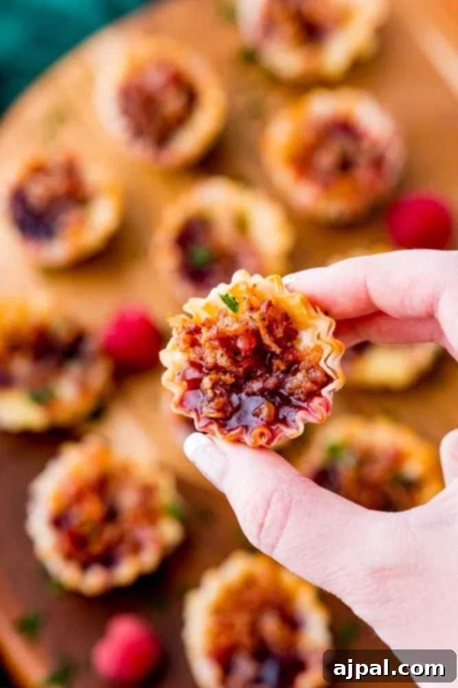 Cheesy Bacon Raspberry Morsels 14 A hand gently lifts a single, perfect phyllo cup, showcasing the harmonious blend of creamy cheese, sweet raspberry jam, and crunchy bacon bits on a wooden board.