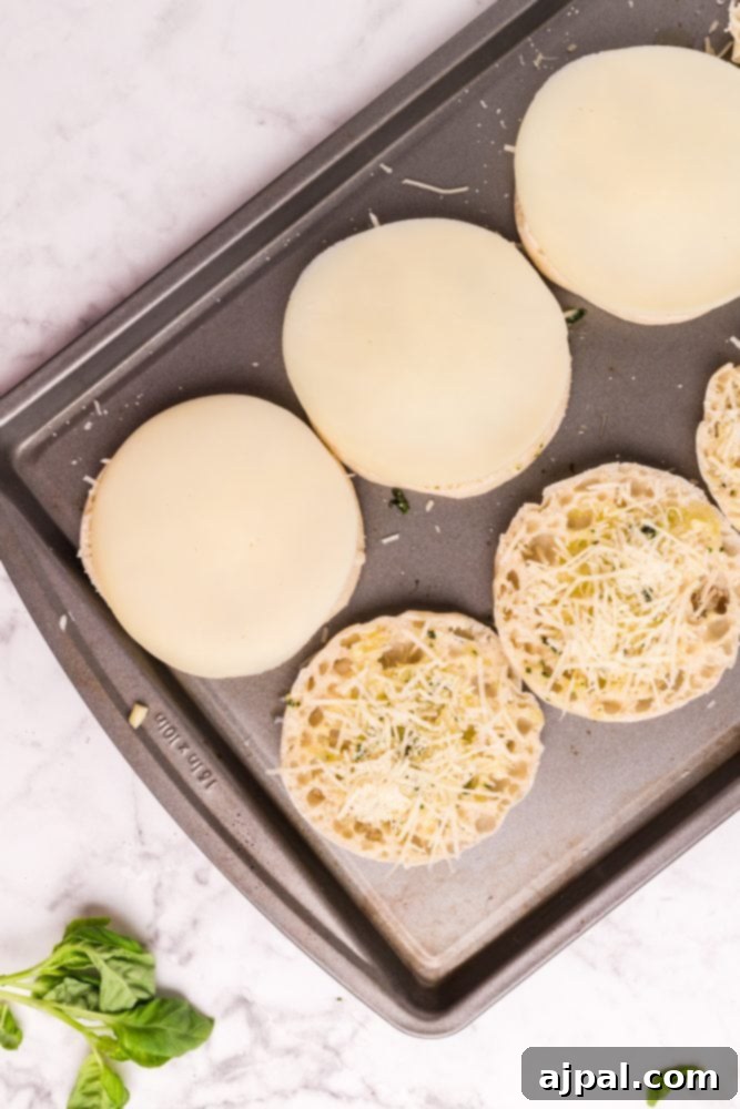 English muffins on a baking sheet, some topped with cheese, ready to be toasted.
