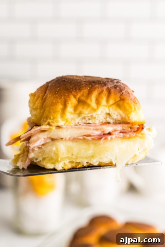 A baked ham and cheese slider held aloft on a spatula, showcasing its melted cheese and golden bun.