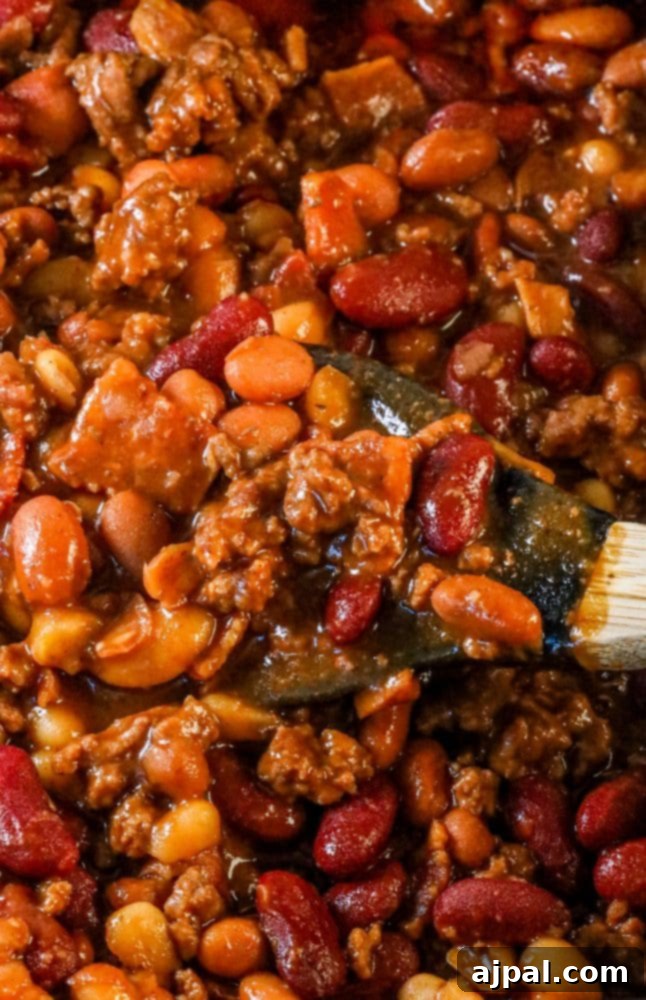 A bowl of the best ever BBQ baked beans, generously loaded with beef and bacon, served with a wooden spoon for easy scooping.