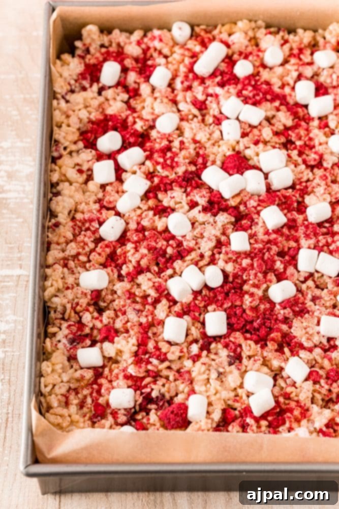 White Chocolate Raspberry Krispie Delights 12 Rice Krispie treats being pressed into a pan, with visible raspberries and marshmallows mixed in.
