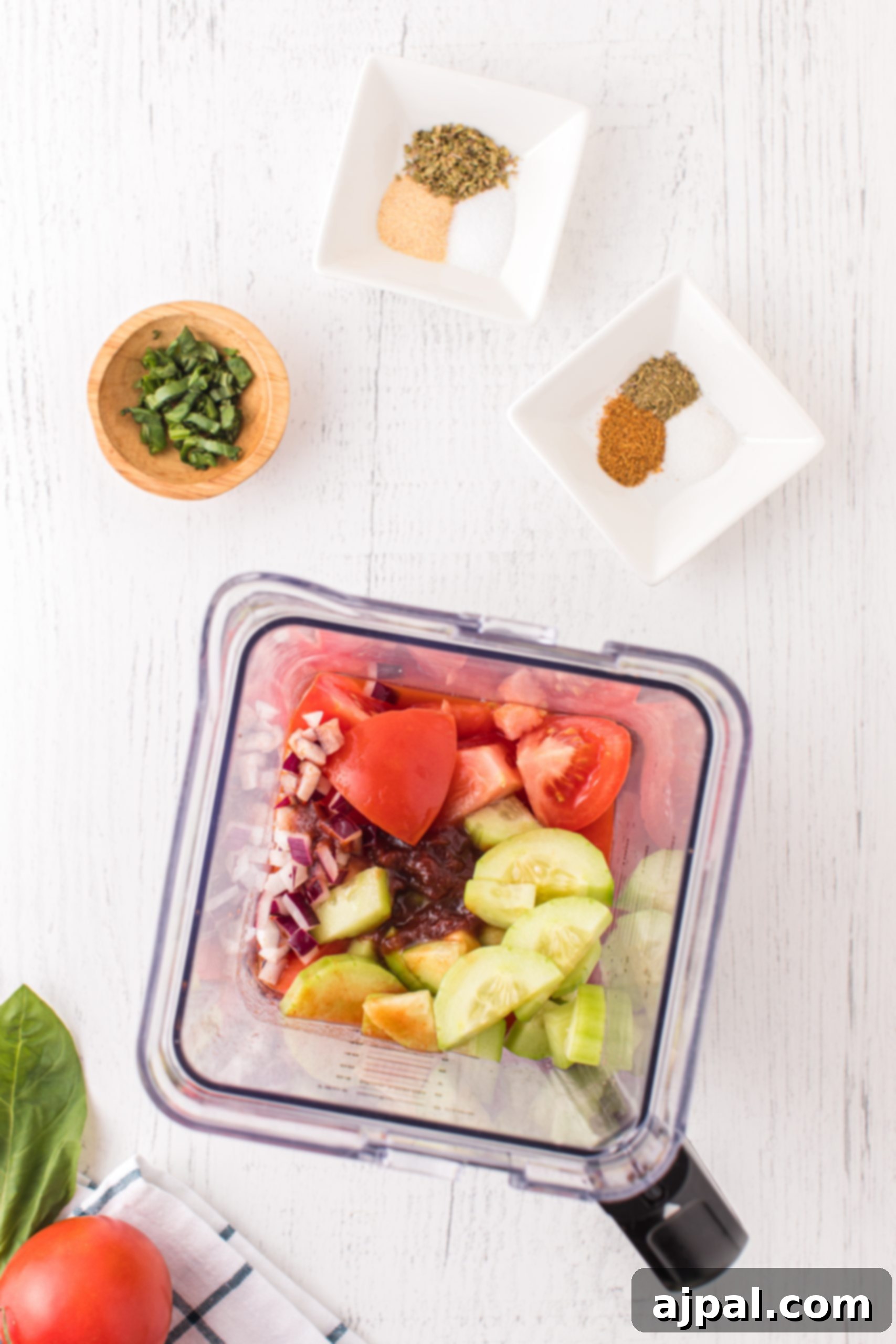 A wooden table showcasing a blender filled with fresh vegetables and spices, ready for making gazpacho.