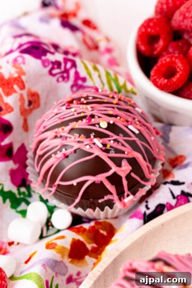 Berry Burst Hot Cocoa Spheres 2 Raspberry hot chocolate bombs topped with chocolate-covered marshmallows on a floral tablecloth.