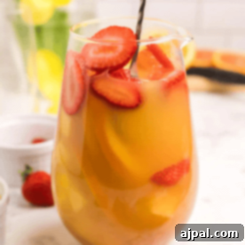 A glass pitcher filled with a refreshing Easy Sangria Mimosas recipe—orange juice, sliced strawberries, and other fruits—graces the marble countertop. A black spoon is submerged in the vibrant drink, inviting you to indulge.