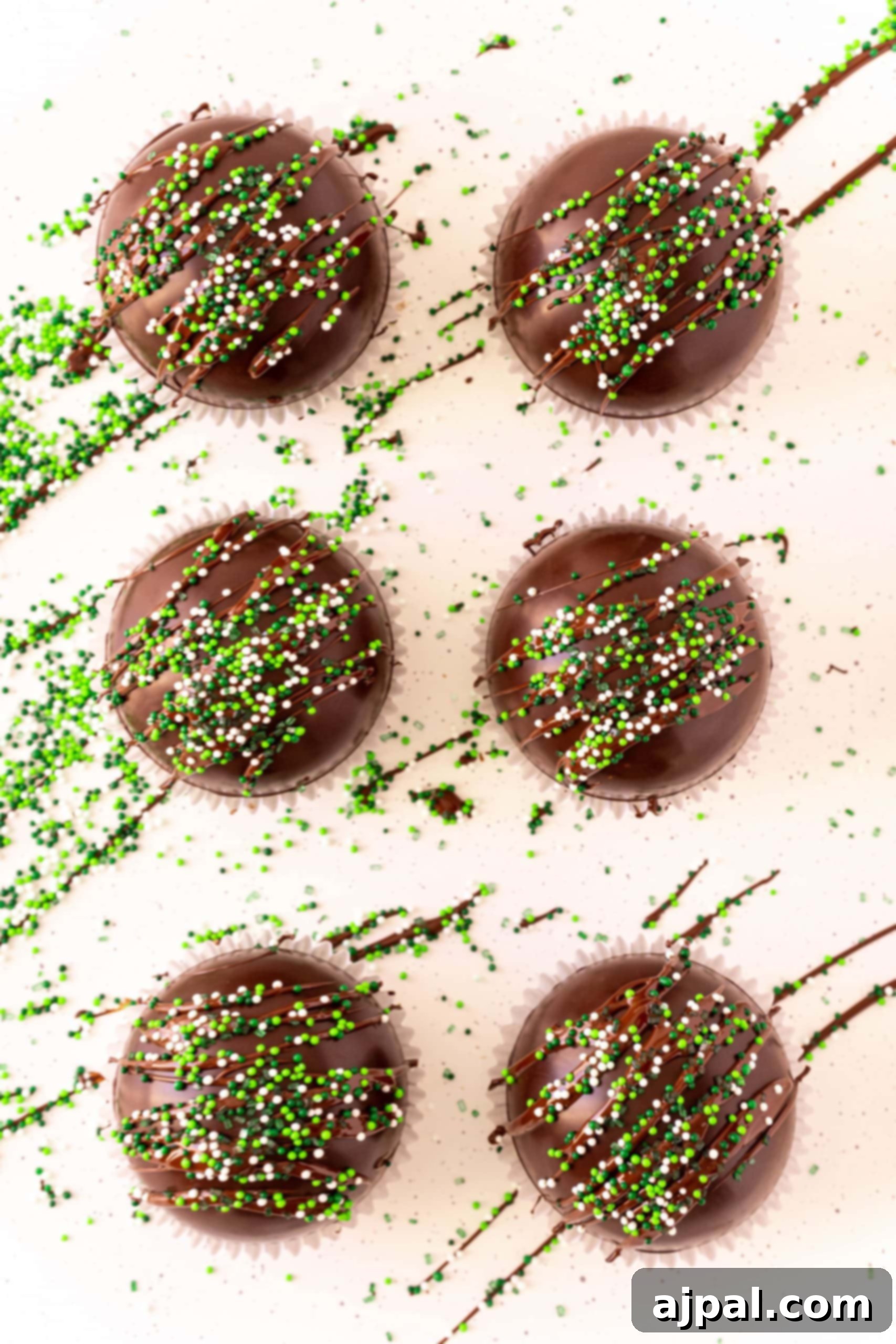 Irresistible Mint Hot Chocolate Bombs 10 chocolate spheres on a white background drizzled with chocolate and covered in green sprinkles.