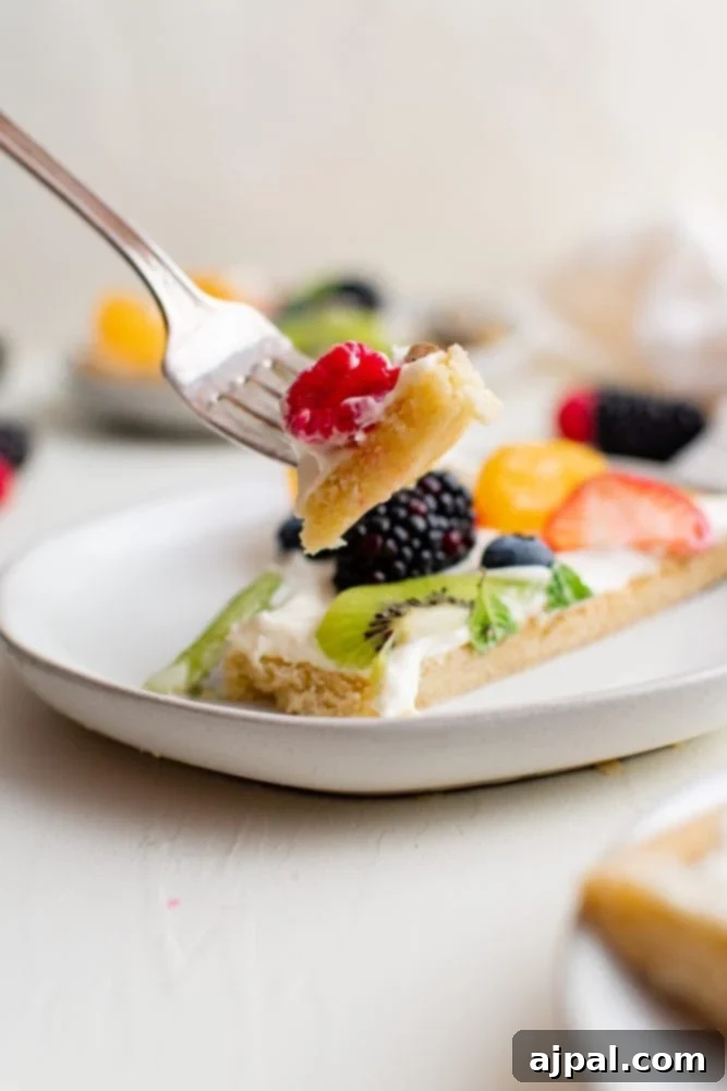 Effortless Rainbow Fruit Pizza 13 A fork lifts a slice of fruit pizza adorned with colorful fruits and creamy topping, showcasing its delicious layers.