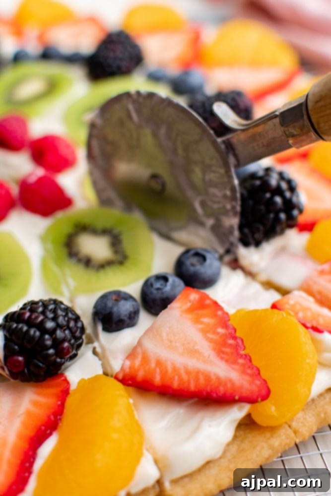Effortless Fruit Pizza Perfection 17 A pizza cutter expertly slicing through a fresh fruit pizza, revealing its layers of cookie, cream, and colorful fruit.