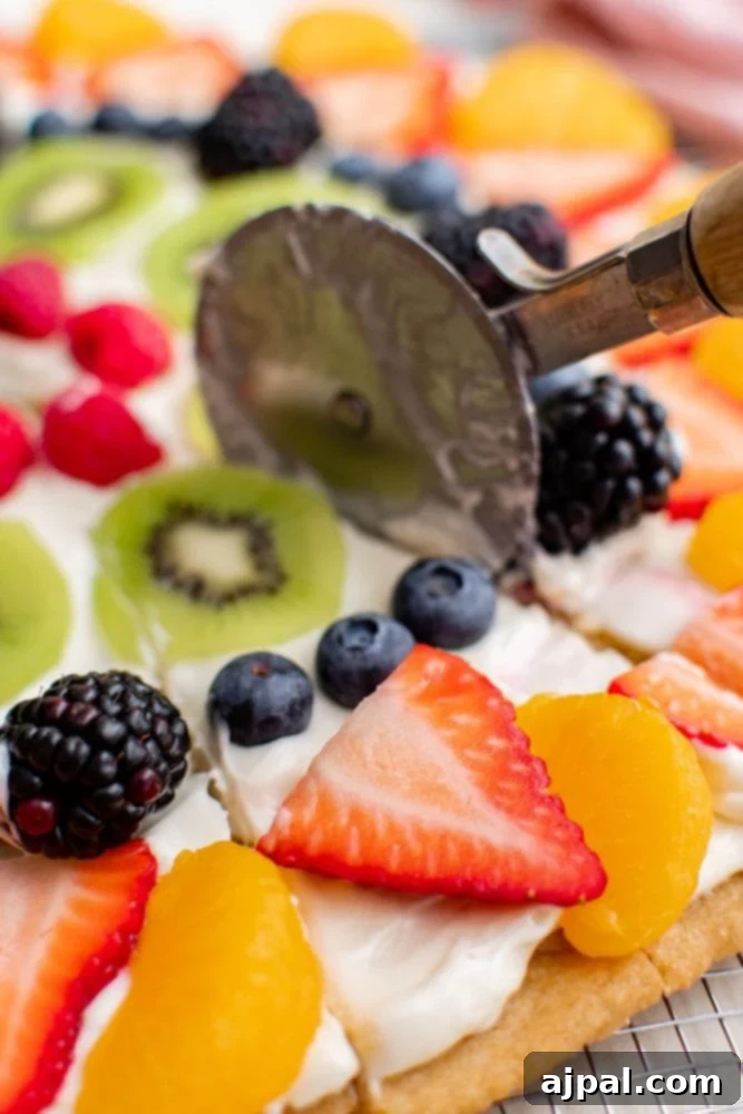 Effortless Rainbow Fruit Pizza 16 A fruit pizza being cut into slices with a pizza cutter, showing the clean layers of crust, cream, and fruit.