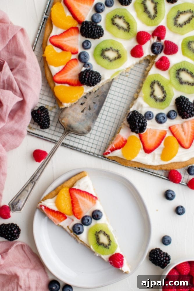 Effortless Rainbow Fruit Pizza 8 A whole fruit pizza with a slice removed, displaying the layers of cookie, cream cheese frosting, and colorful fruit toppings.