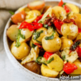 A vibrant bowl of vegan Italian potato salad, featuring tender baby potatoes, bright red bell peppers, tangy artichoke hearts, fragrant herbs, and sweet sun-dried tomatoes, all tossed in a light oil-based dressing.