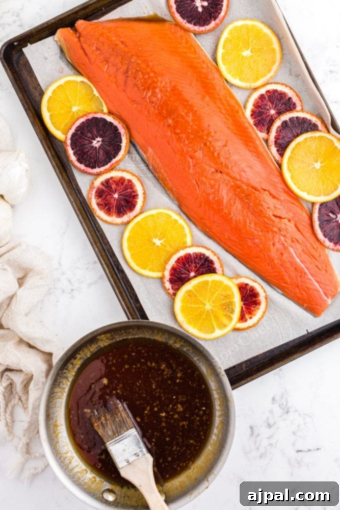 A pan of rich orange glaze sits next to a raw salmon fillet on a foil-lined baking sheet, accompanied by fresh orange slices, ready for the oven.