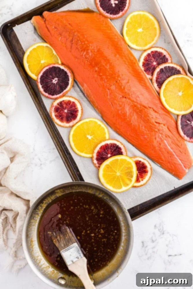 A pan of rich orange glaze sits next to a raw salmon fillet arranged on a foil-lined baking sheet, accompanied by fresh orange slices, ready for baking.