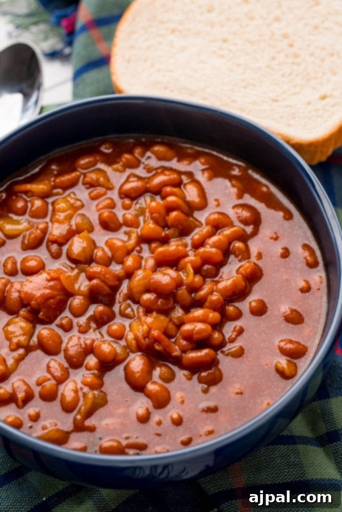 A rustic image of slow cooker baked beans served in a blue bowl, accompanied by a slice of bread on a plaid tablecloth.