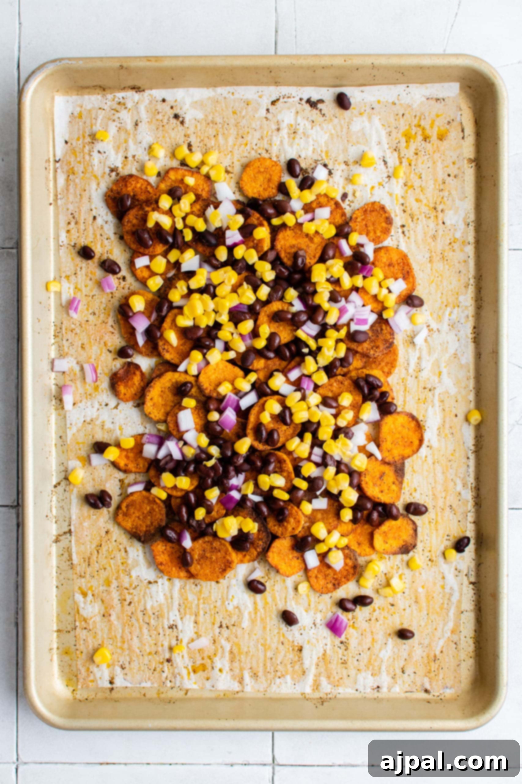 A sheet pan with sweet potato chips, partially covered with the first layer of savory nacho toppings like beans and corn.