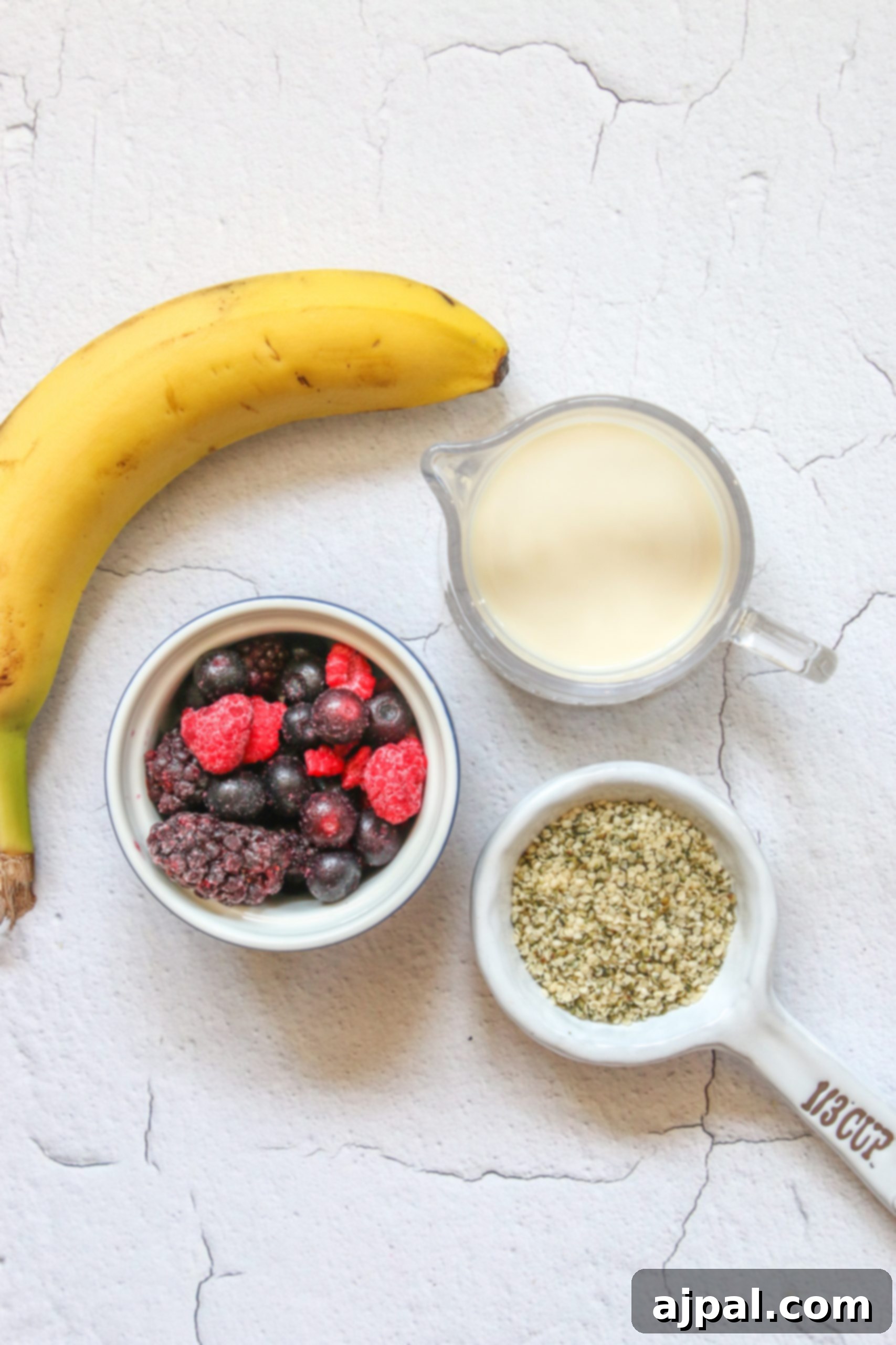 Triple Berry Smoothie Bowls 6 An array of ingredients for a triple berry smoothie bowl laid out on a table: a banana, mixed berries, almond milk, and hemp hearts, all ready for blending.