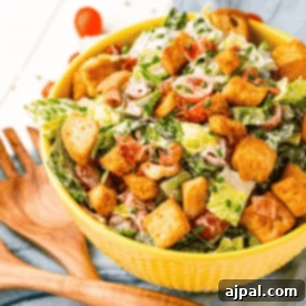 Effortless BLT Salad 8 A yellow bowl filled with a BLT salad featuring croutons, cherry tomatoes, and bacon bits is placed on a blue cloth next to wooden salad utensils.