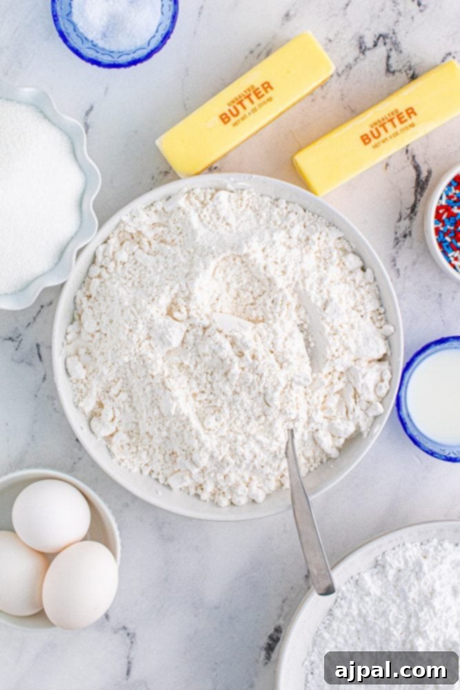 Stars and Stripes Sugar Cookie Bars 7 Baking ingredients for sugar cookies, including flour, eggs, butter, and sugar, artfully arranged on a wooden surface.