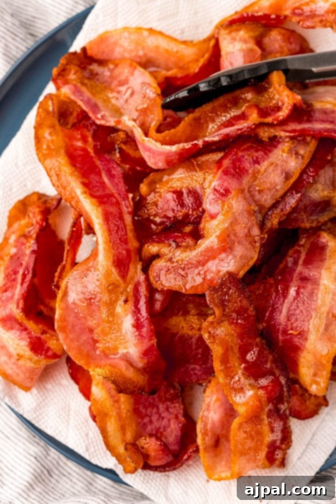 Tongs lifting a strip of perfectly cooked bacon onto a plate lined with paper towels.