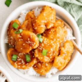 A bowl of chicken breast sesame chicken served over white rice, garnished with green onions and sesame seeds.