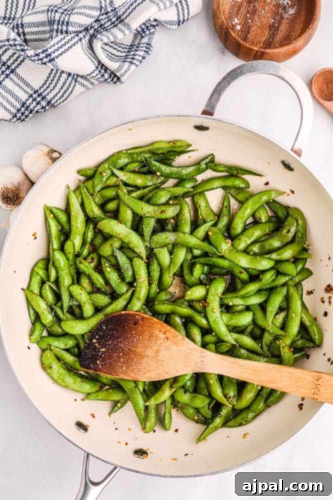 Spicy green edamame beans are cooked in a skillet with a wooden spoon, showcasing the rich seasoning.