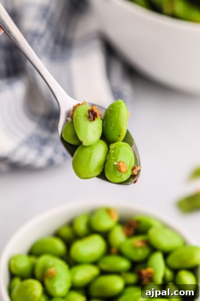 A person holds a spoonful of spicy green edamame beans in a white bowl, ready to serve.
