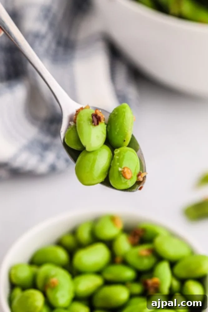 A spoonful of vibrant green, spicy edamame beans is gracefully lifted from a white bowl, showcasing their perfect seasoning and enticing color.
