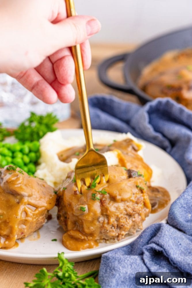 Rich Salisbury Steaks with Velvety Mushroom Gravy 3 A fork piercing a tender Salisbury steak submerged in mushroom gravy, served with creamy mashed potatoes.