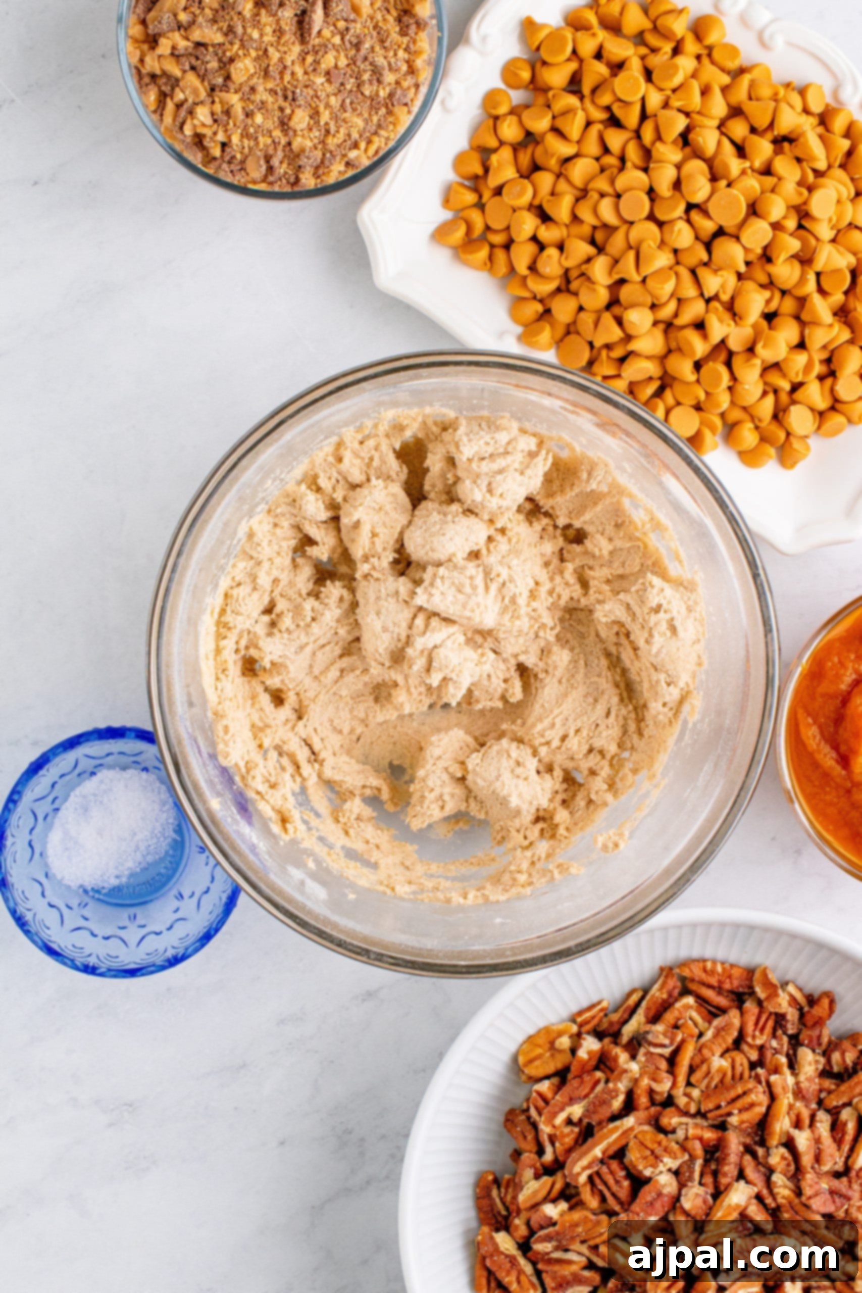 Cookie dough being blended in a glass bowl, with butterscotch chips and nuts ready on a table
