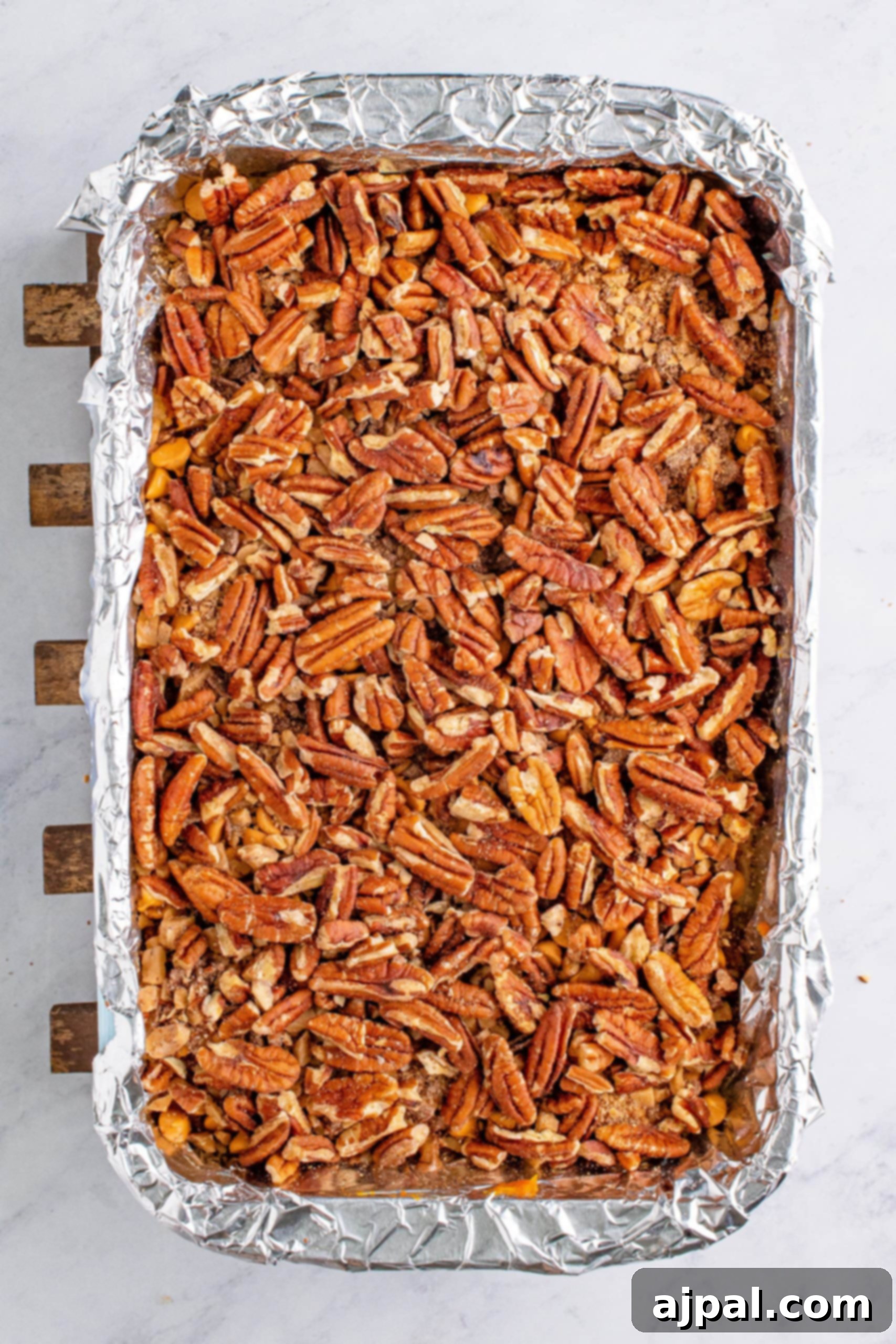 A cookie base topped with pumpkin filling and a generous layer of pecans in a baking sheet lined with foil
