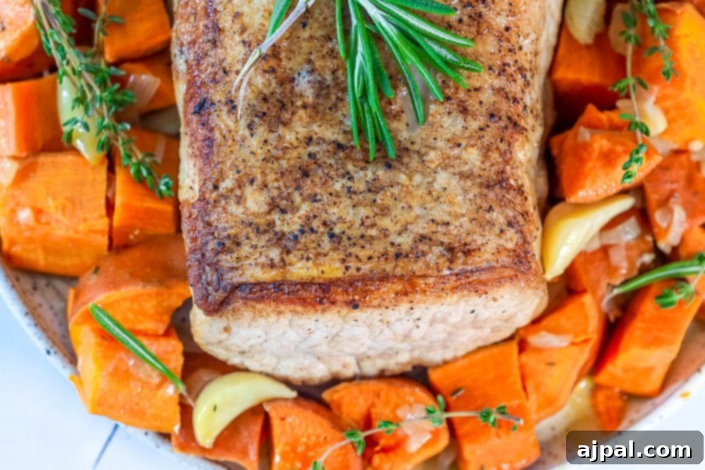 A sheet pan filled with roasted sweet potatoes, garlic, and onions, with a perfectly cooked pork loin nestled among them, garnished with fresh rosemary.