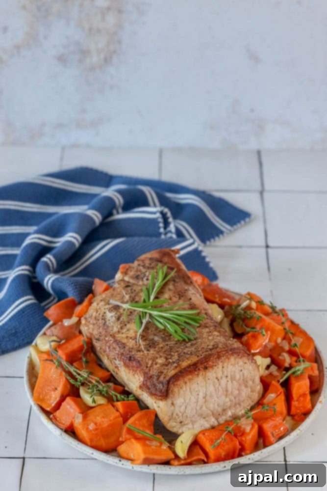 A close-up of a serving of roasted pork loin, sweet potatoes, and rosemary on a rustic white plate, highlighting the golden-brown crust of the pork.