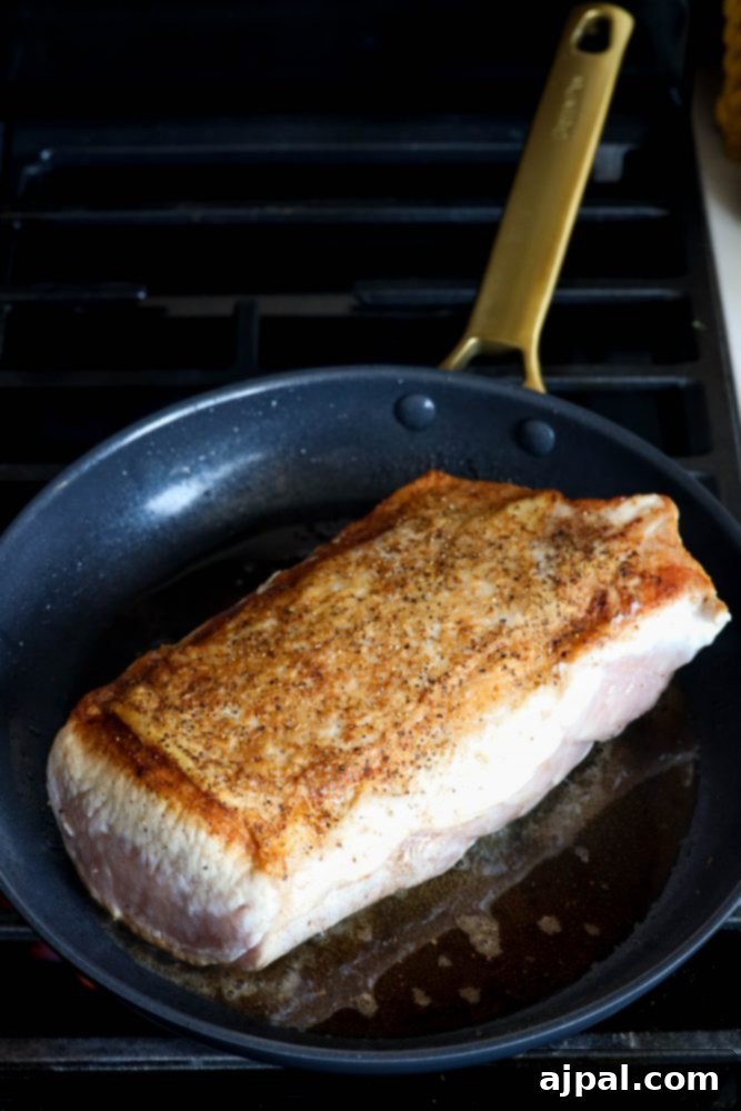 A pork loin being seared to a golden brown in a hot pan, preparing it for roasting.