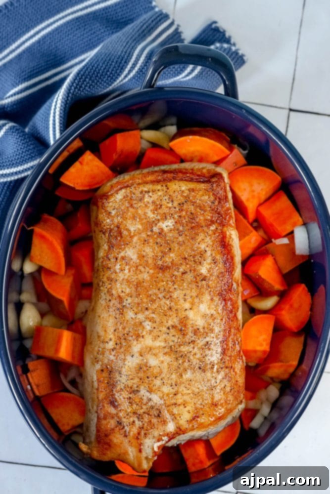 The seared pork loin placed on top of sweet potatoes and herbs in a roasting pan, ready for the oven.