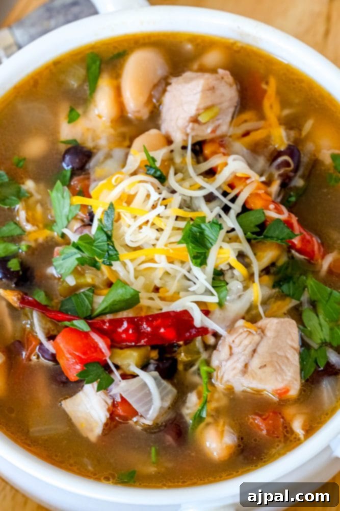A steaming white bowl of hearty Green Chile Chicken Soup, garnished with vibrant cilantro, shredded Monterey Jack cheese, and a hint of red chili flakes on top of tender chicken, peppers, and beans.