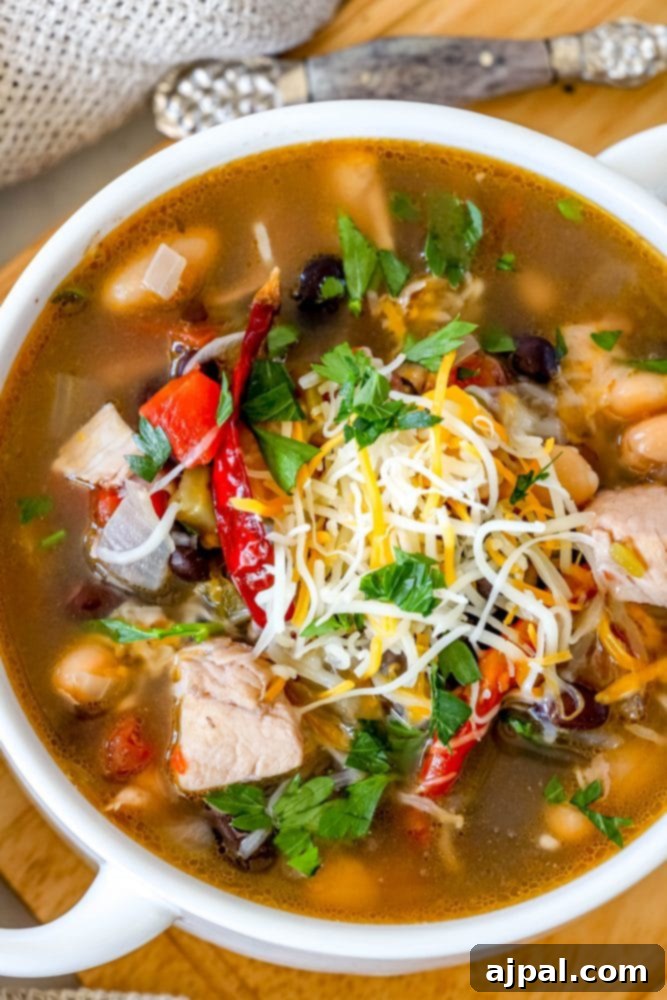 A close-up shot of a white bowl filled with creamy Green Chile Chicken Soup, highlighting the rich broth, tender chicken, and various beans.