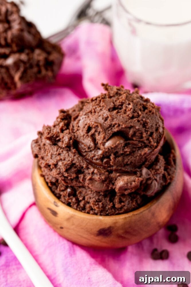 No-Bake Brownie Batter 2 A close-up of delicious, fudgy edible brownie batter in a rustic wooden bowl, ready to be enjoyed. The batter is rich brown, suggesting deep chocolate flavor, and sits on a soft pink cloth.