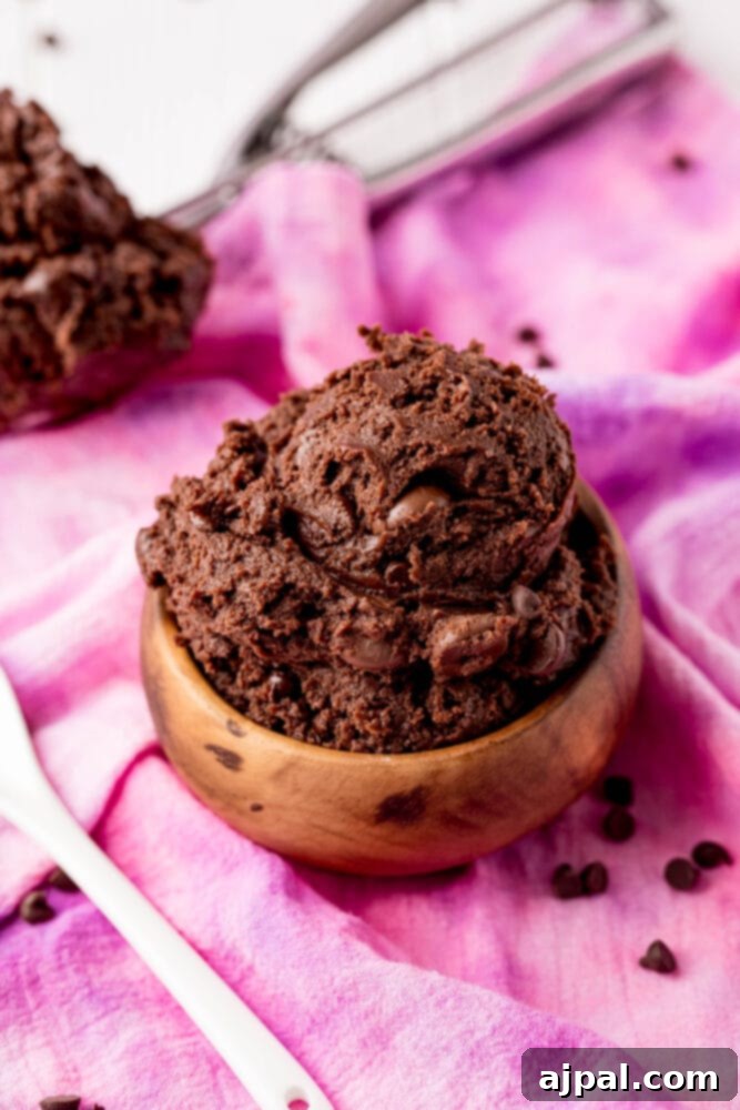 No-Bake Brownie Batter 9 A generous serving of decadent edible brownie batter in a rustic wooden bowl, with a spoon resting in it, inviting a taste of its rich, fudgy texture. The bowl sits on a soft pink cloth.
